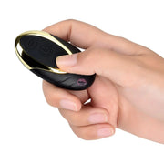 A hand holds The Secret Affaire’s Mr. Lover Sucking Thrusting Dildo Vibrator, a small oval-shaped black and gold device with two buttons visible on its surface.